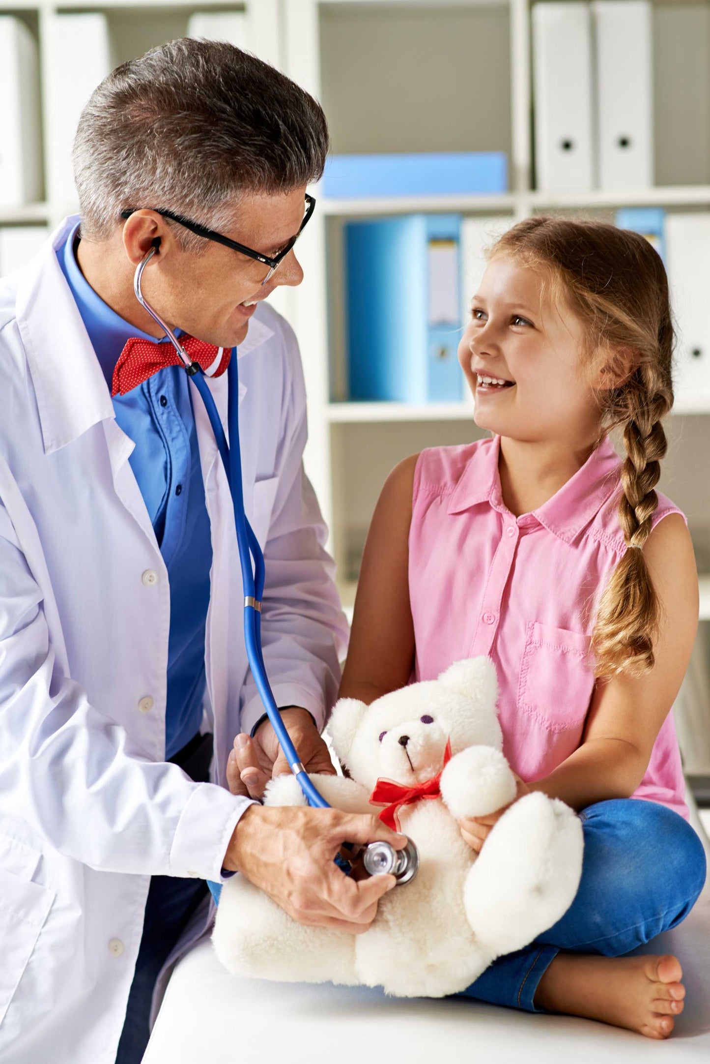 Pediatrician using stethoscope on a smiling young girl holding a teddy bear during a pediatric consultation